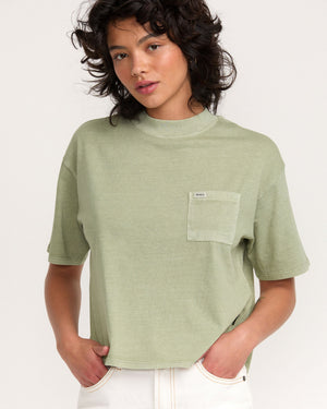 A person with wavy dark hair wears the Kinney Tee Pocket PTC in light green and off-white pants, standing against a plain light background.