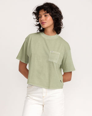 A person with short, curly dark hair wears the loose-fitting Kinney Tee Pocket PTC in light green and white high-waisted pants, standing against a plain light background.