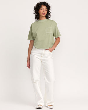 A person with curly hair wears the Kinney Tee Pocket PTC, a loose, light green short-sleeve top with a chest pocket, paired with white wide-leg pants against a plain white background.